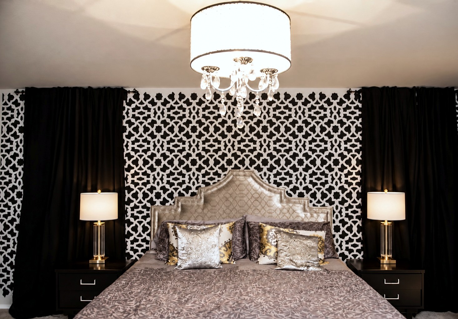 Bedroom with a black and white stenciled accent wall, a nailhead headboard, matching lamps and a crystal chandelier