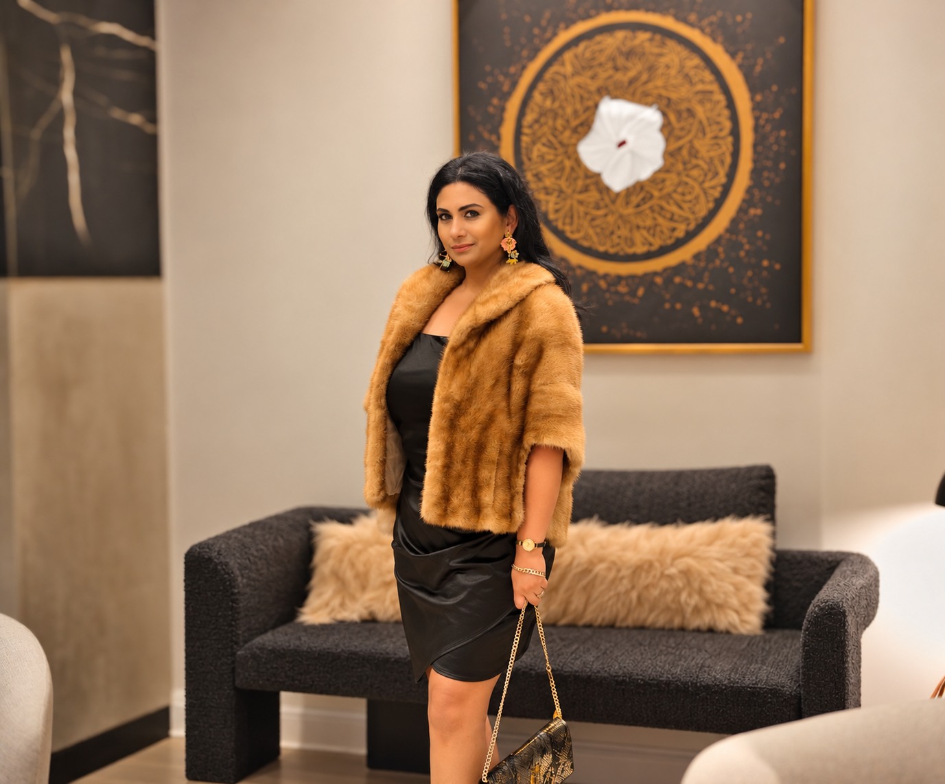 Founder in a black satin dress and a tan fur stole beside a dark boucle sofa